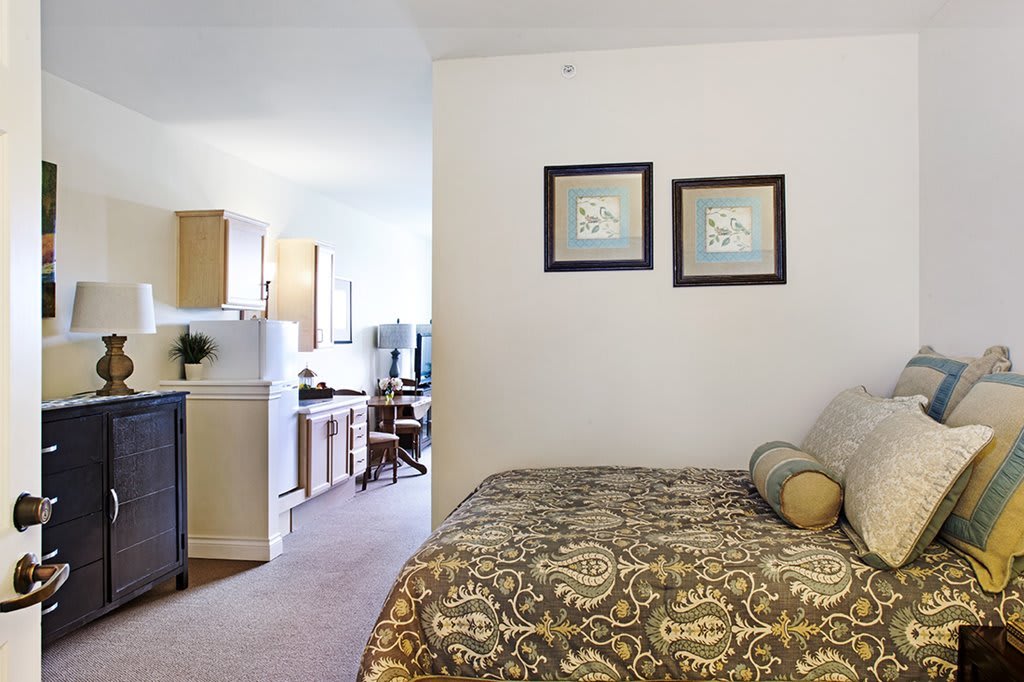 A cozy apartment bedroom with a bed and a small kitchen area in the background.