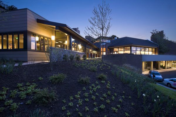 A modern building with illuminated exteriors at dusk, featuring landscaped surroundings.