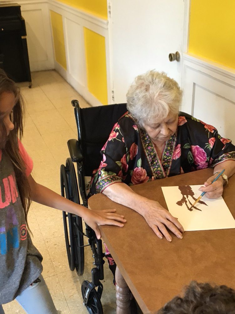 An elderly woman in a wheelchair engages in drawing with a young girl observing.