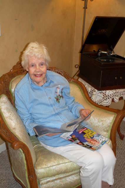 An elderly woman smiles while sitting in a vintage armchair, holding a magazine in a cozy living room.