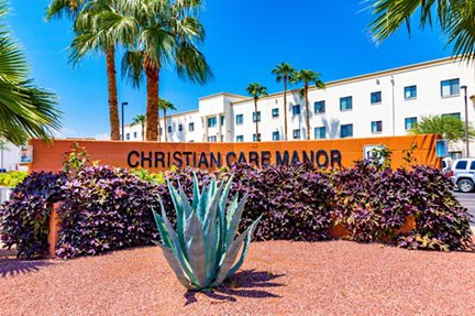 The entrance to Christian Care Manor with palm trees and a succulent in the foreground under a clear blue sky.