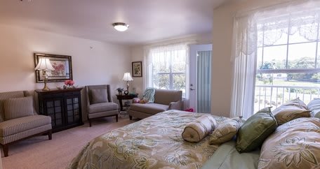 A cozy bedroom with a bed, seating area, and natural light streaming through the windows.
