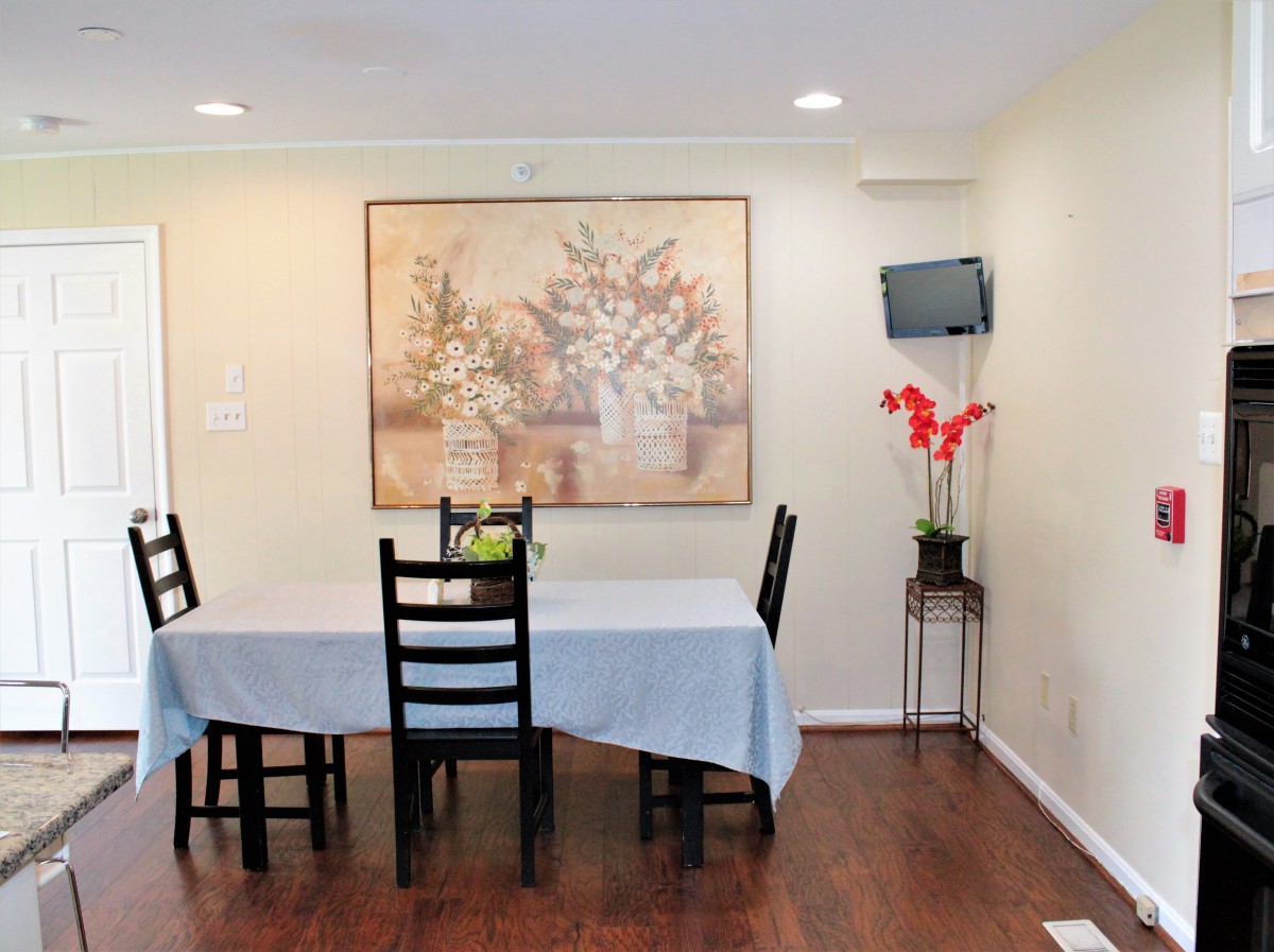 A cozy dining room with a table set, wall art, and a small television mounted beside a flower vase.
