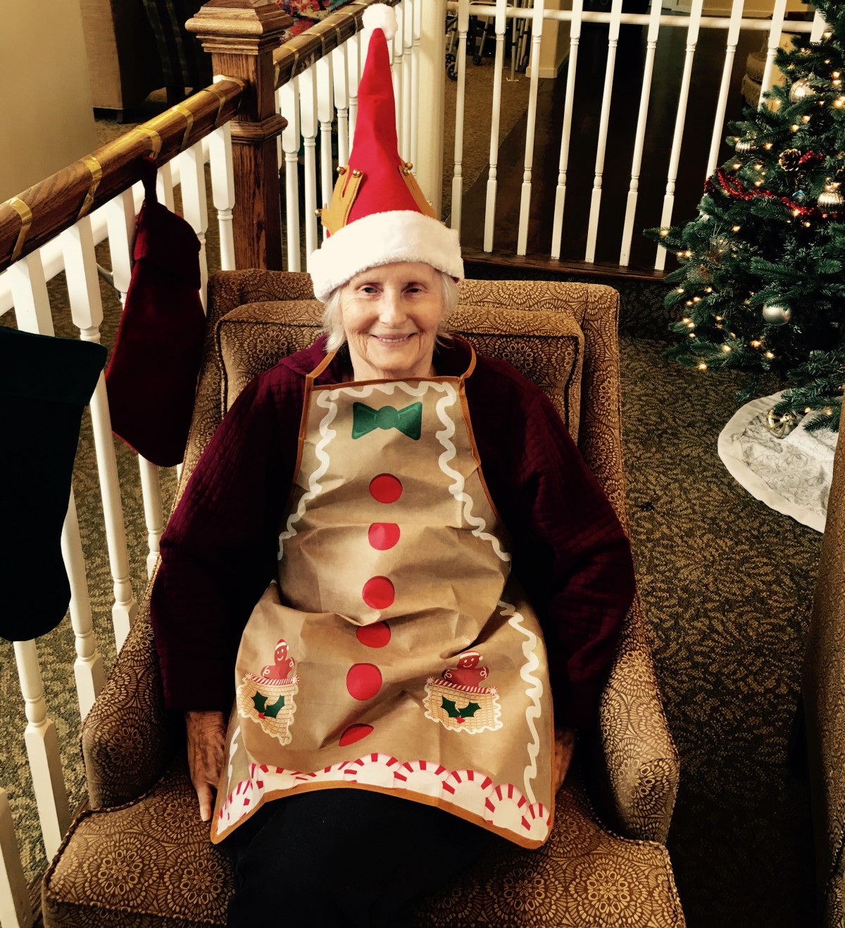A person wearing a Santa hat and festive apron sits in a chair next to a Christmas tree.