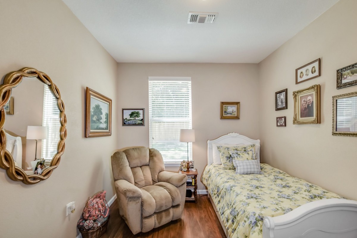 A cozy bedroom featuring a single bed with floral bedding, a reclining chair, and various artworks on the walls.