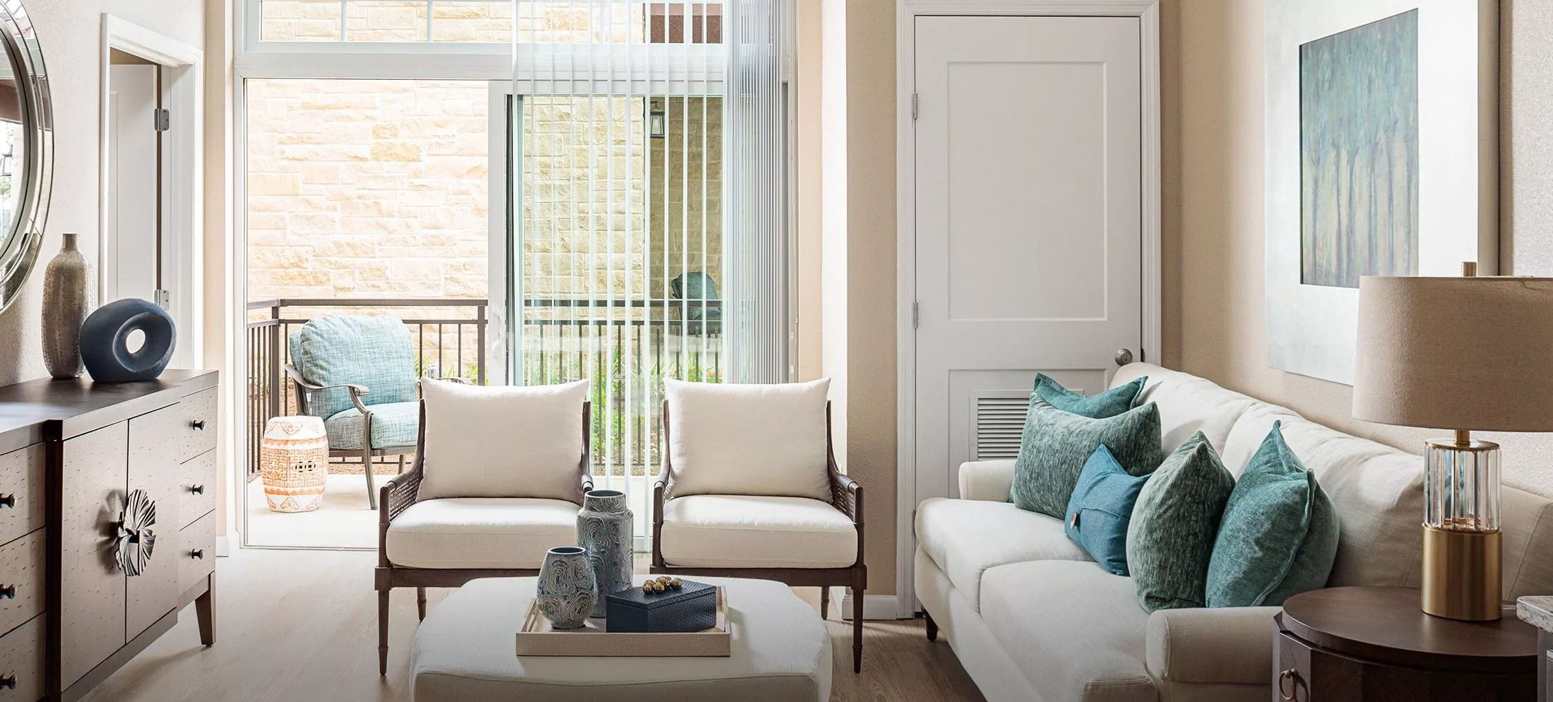 A cozy living room with a white sofa, two chairs, and a coffee table decorated with vases, set beside large windows with blinds.