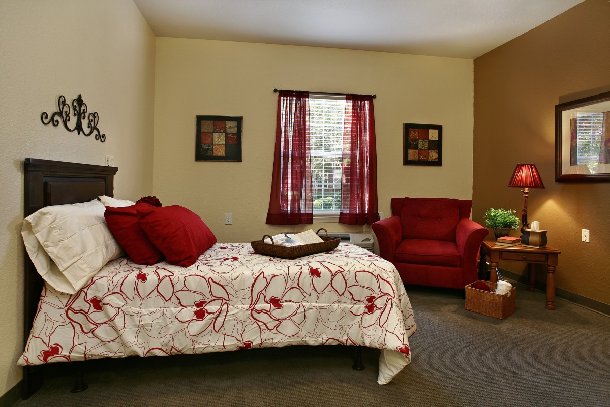 A cozy bedroom with a bed featuring floral bedding, a matching red chair, and decorative curtains.