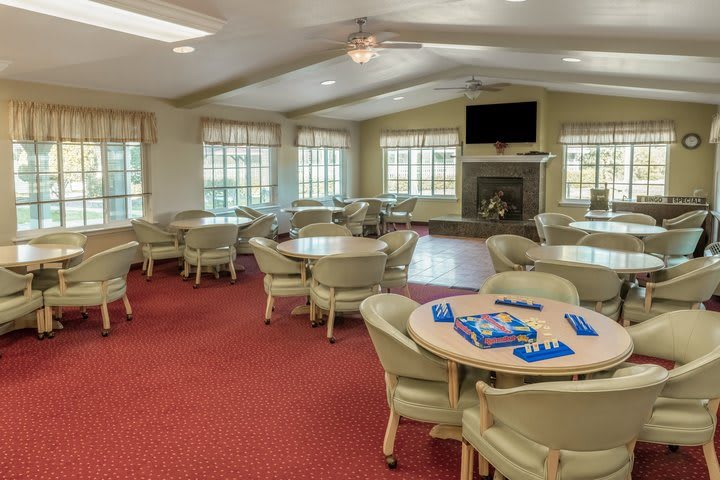 A spacious community room with tables and chairs set up for games, featuring large windows and a television.