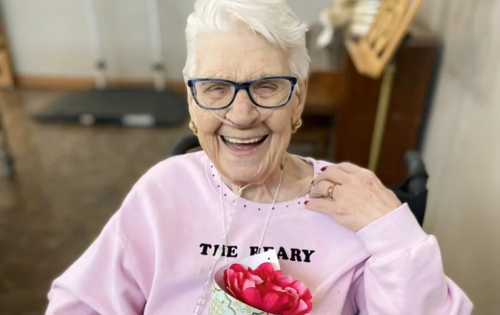 An elderly woman with glasses smiles while holding a gift indoors.