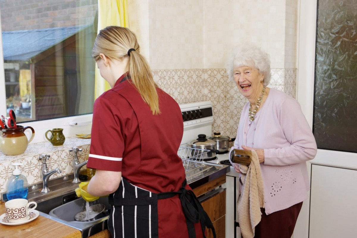 A caregiver assists an elderly woman in the kitchen.