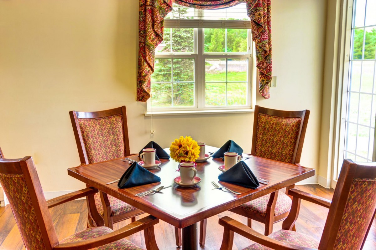 A cozy dining area with a wooden table set for four, featuring flowers and napkins, near a window with a view.