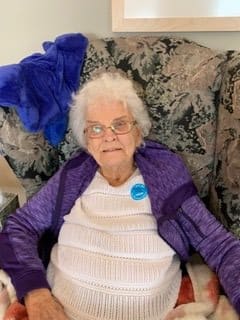 An elderly woman wearing glasses and a purple shawl is seated on a sofa.
