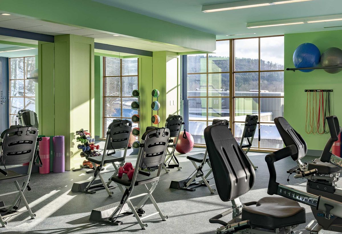 A brightly lit gym with various workout equipment and benches, featuring large windows allowing natural light in.