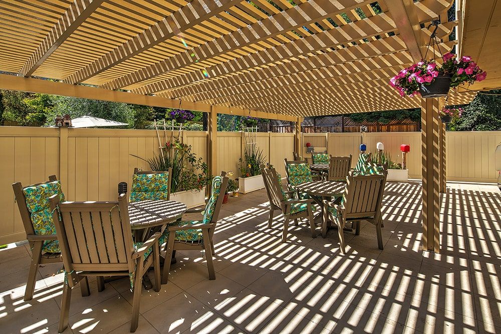 A charming outdoor patio area featuring a pergola with wooden slats creating patterned shadows, surrounded by lush garden plants and complete with elegant patio furniture.