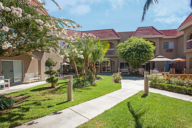 A beautifully landscaped courtyard with a central pathway surrounded by lush greenery and trees, adjacent to a building under a clear blue sky.
