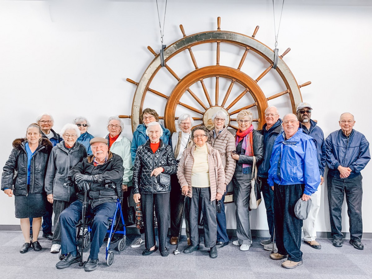 A group of senior individuals standing in front of a large decorative wheel indoors.