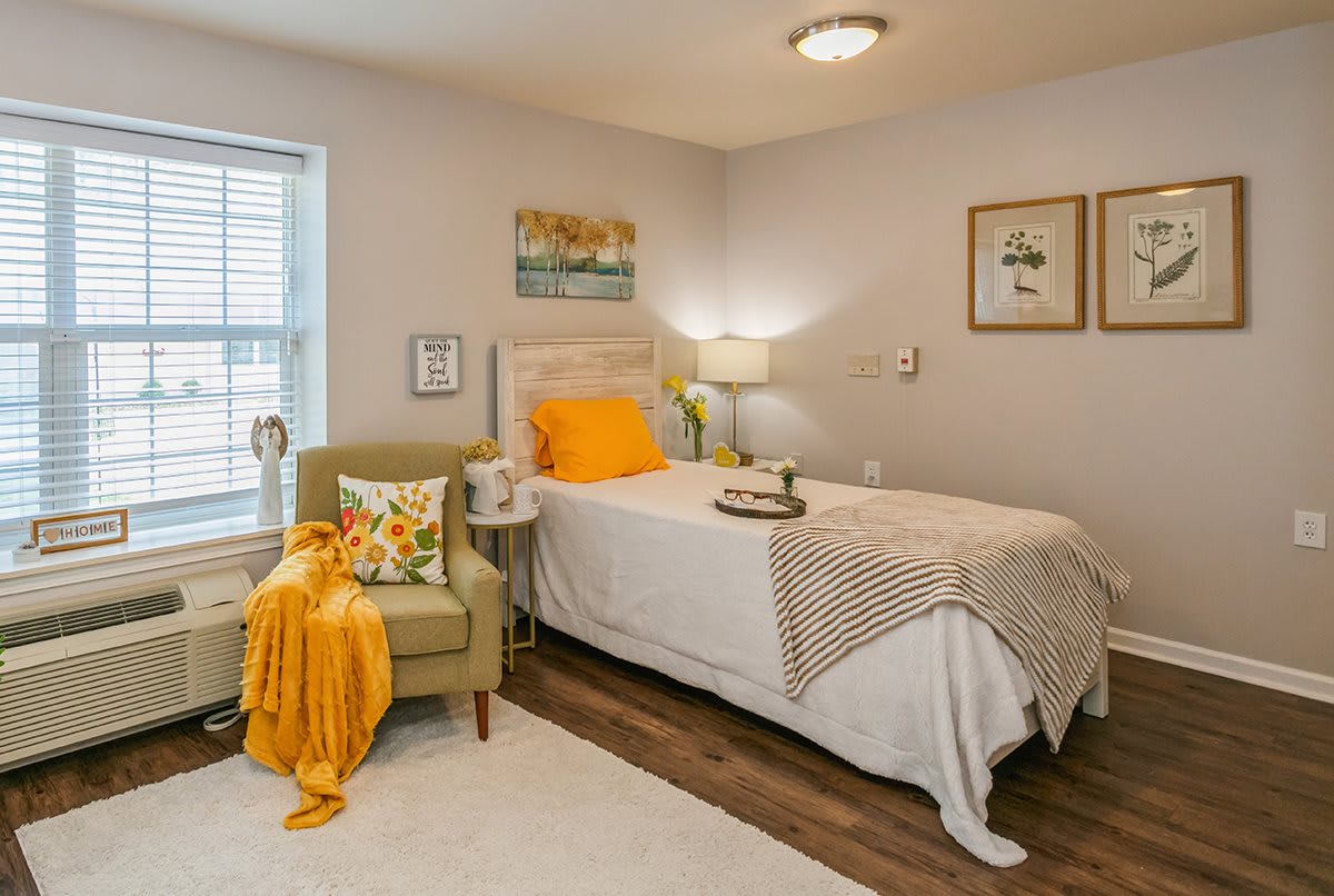 A cozy bedroom with a single bed, decorated in warm tones and featuring an armchair, artwork, and a lamp.
