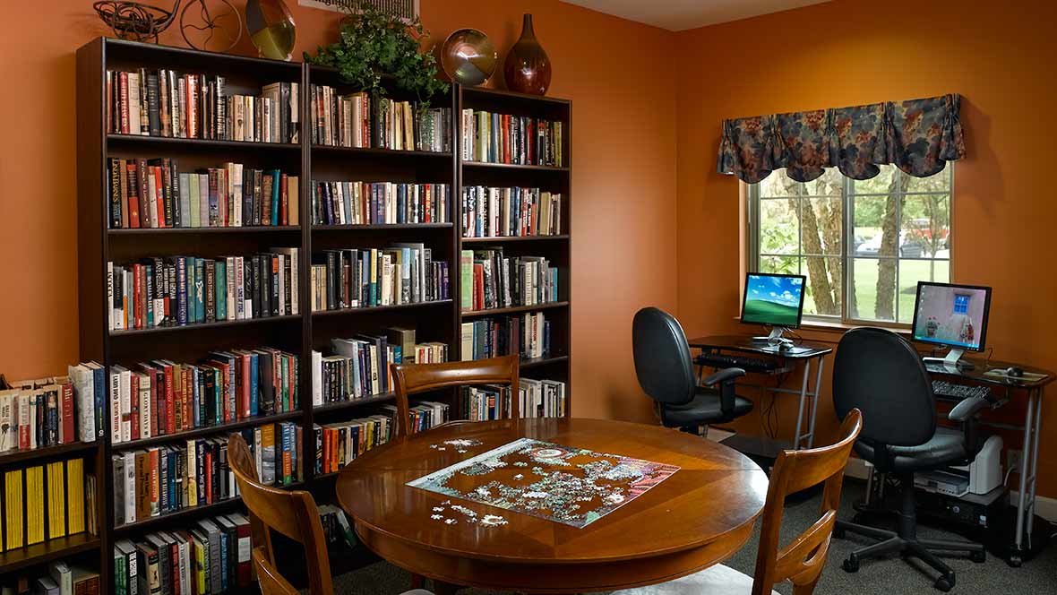 A cozy home library with bookshelves, a table with a puzzle, and a computer by the window.