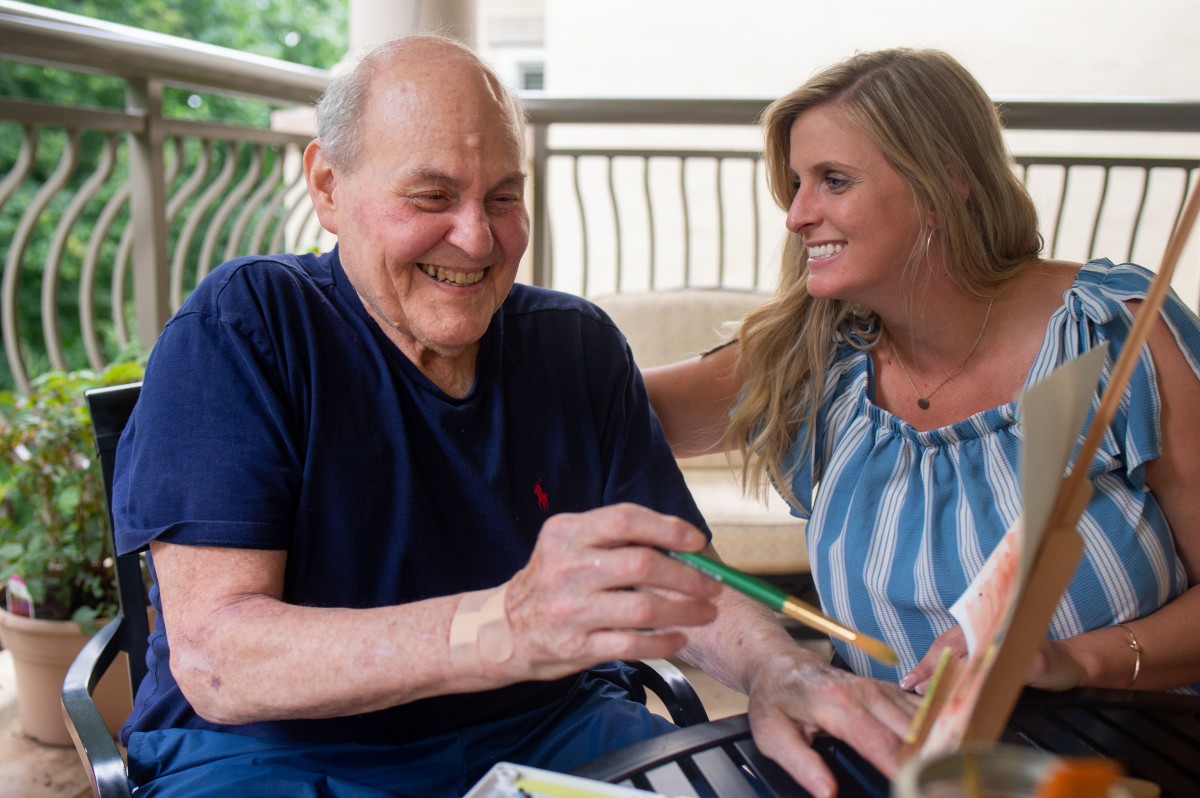 An elderly man and a woman share a joyful moment while painting together outside.