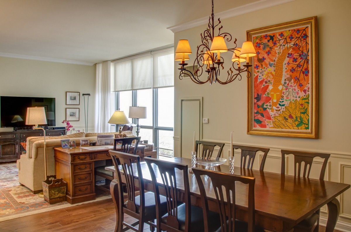 A spacious dining room with a wooden table and chairs, illuminated by an ornate chandelier. A colorful painting decorates the wall.
