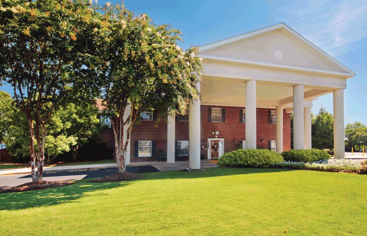 A brick building with white columns on a sunny day, surrounded by a green lawn and trees.