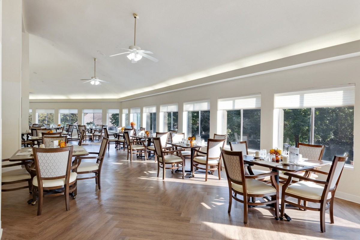 A spacious and bright dining room with multiple tables and chairs, featuring large windows and ceiling fans.