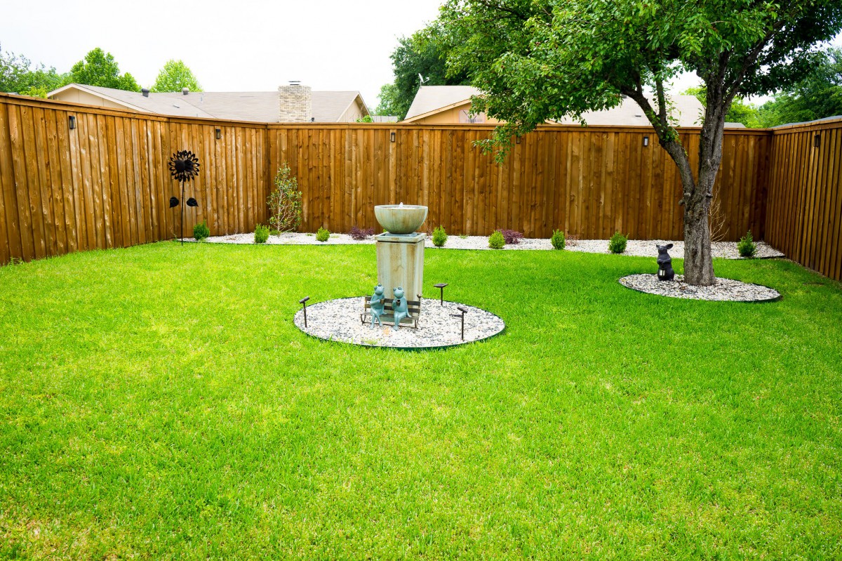 A peaceful backyard garden with a green lawn, wooden fence, and decorative fountain centerpiece.