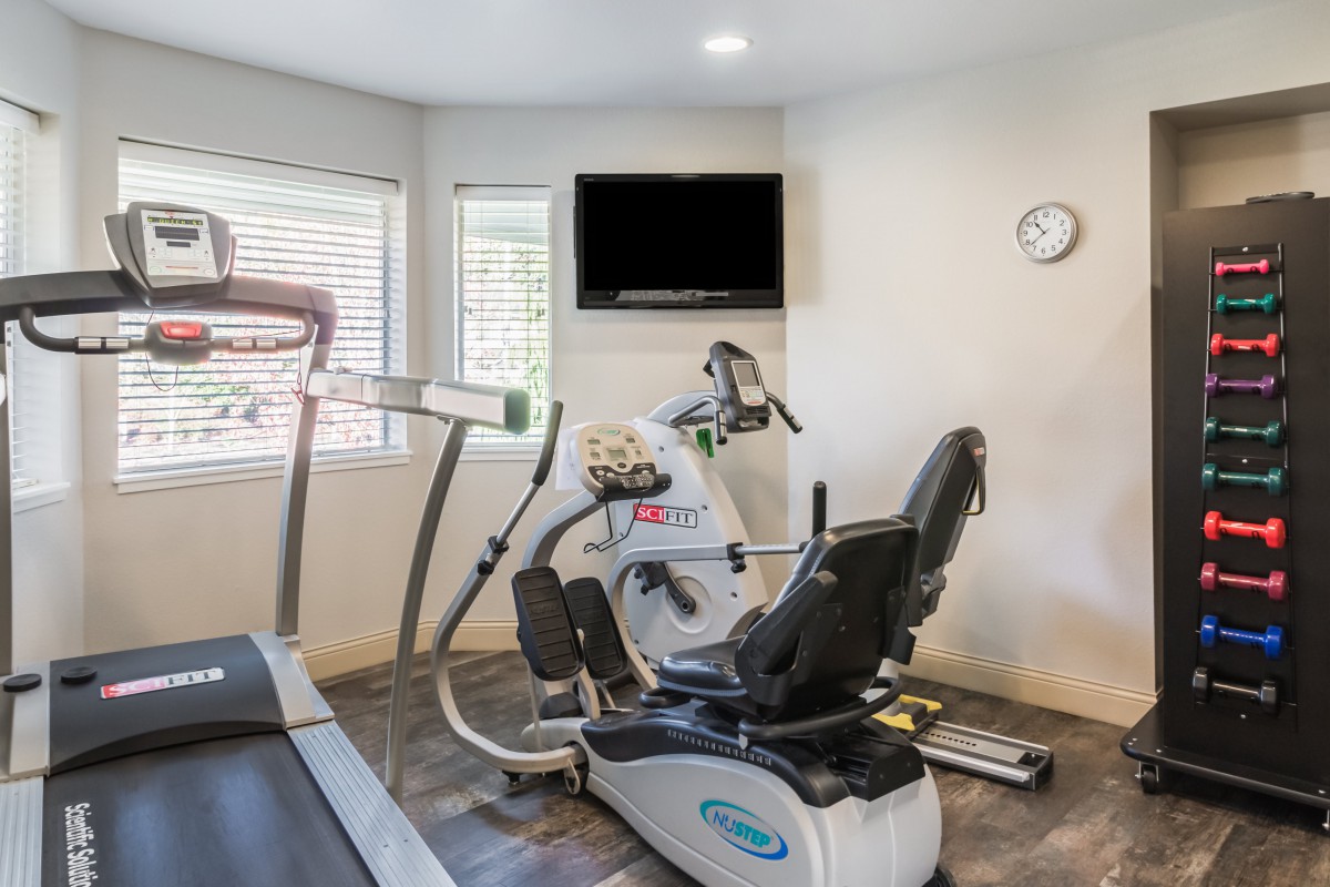 A small fitness room equipped with a treadmill and a recumbent bike, featuring a wall-mounted television and a clock.