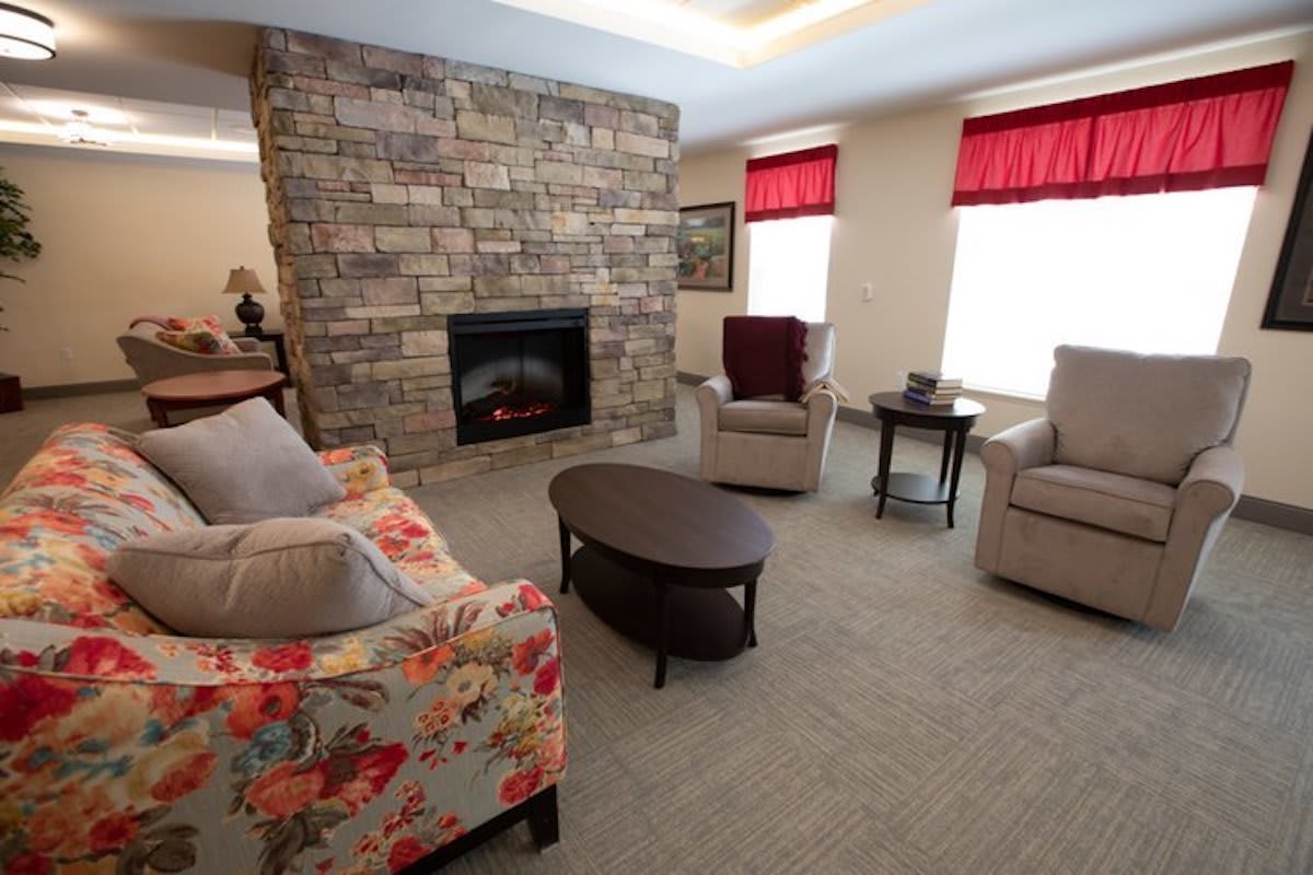 A cozy living room featuring a stone fireplace, comfortable seating, and natural light from windows with red valances.