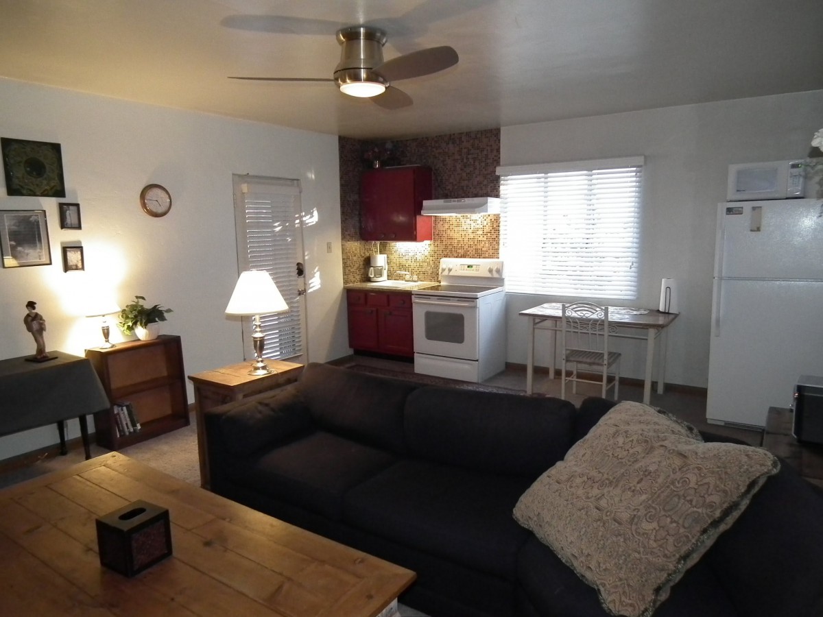 A cozy living room with an open kitchen layout featuring a black sofa, wooden furniture, and a modern ceiling fan.