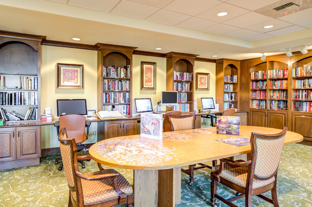 A well-lit library room with bookshelves, computers, and a large table surrounded by chairs.