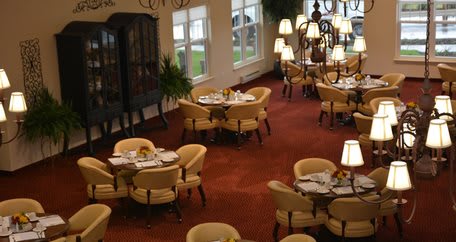 An elegant restaurant dining area with neatly arranged tables and chairs, featuring a warm carpeted floor and ambient lighting.
