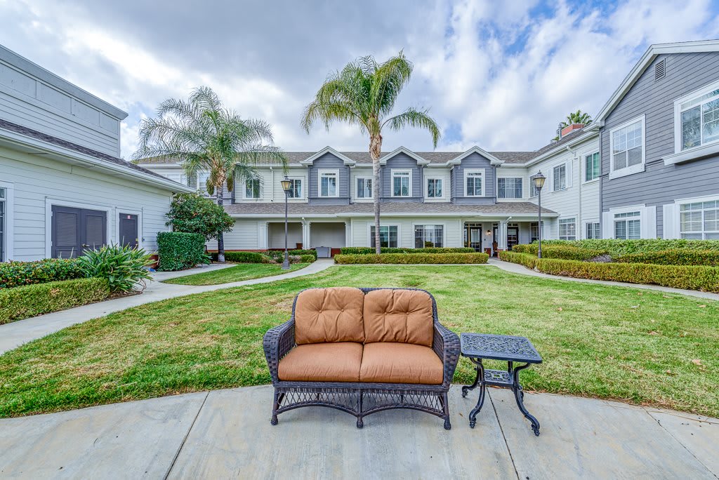 A seating area with a loveseat and table in front of a two-story house with palm trees under a cloudy sky.