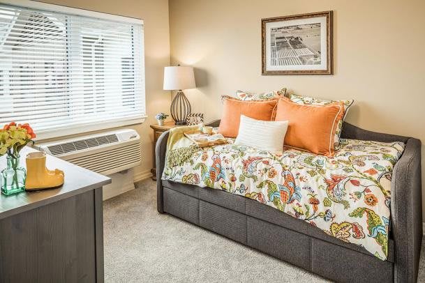 A cozy bedroom with colorful bedding and decorative pillows, featuring a bedside lamp and a window with blinds.