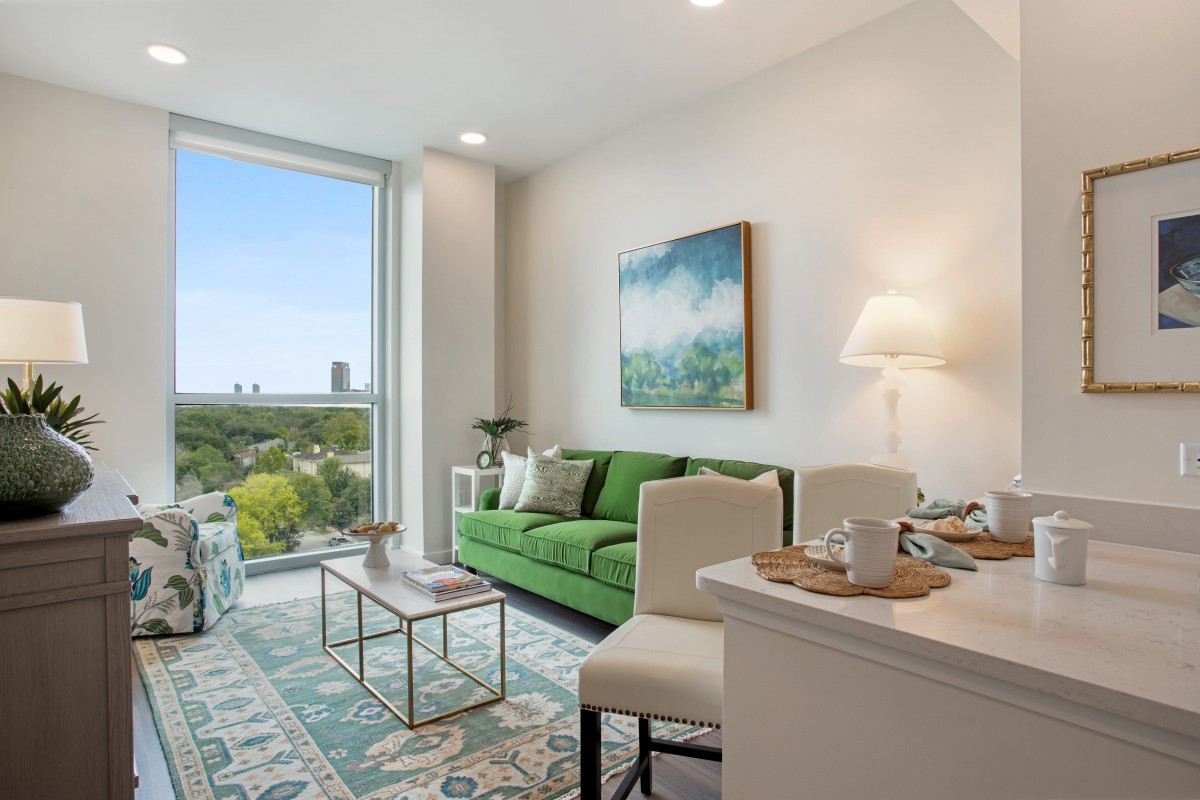 A modern living room with a green couch, coffee table, and large window offering a city view.
