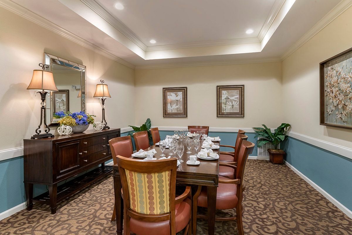 A well-decorated dining room featuring a long table surrounded by chairs, with elegant lighting and decorative elements including paintings, plants, and a large mirror.