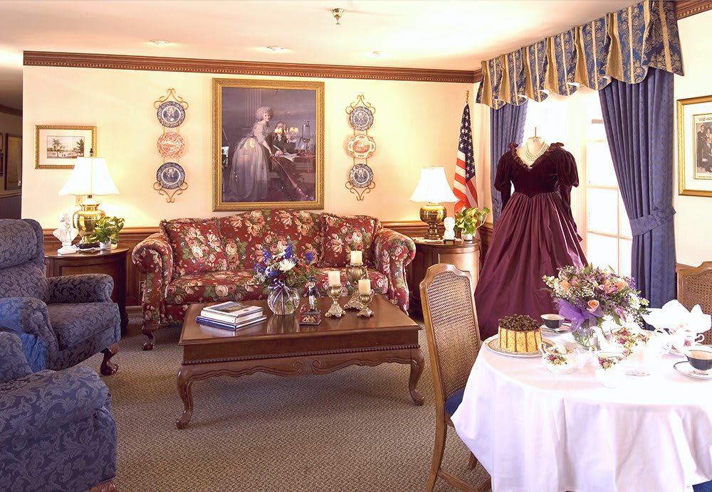 A vintage-themed living room featuring ornate furniture, a historic painting, and a dress on display, with an American flag in the corner.