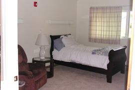 A cozy bedroom featuring a single bed, an armchair, and a bedside lamp, accompanied by a window with curtains.