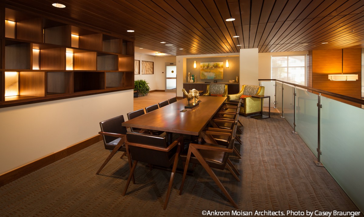 A modern conference room with a large wooden meeting table and comfortable chairs, featuring a warm, wood-paneled ceiling and soft lighting.