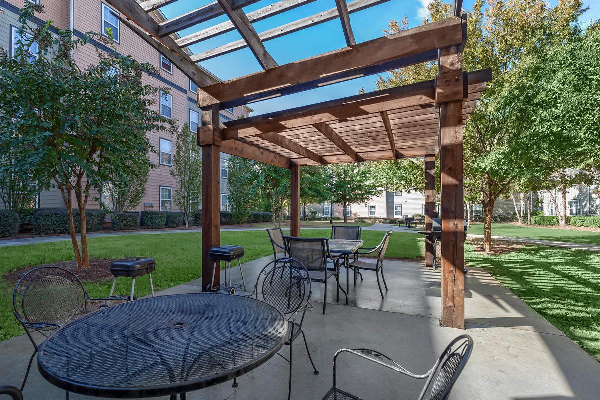 A sunny patio area with a wooden pergola, tables, and chairs surrounded by greenery and buildings.