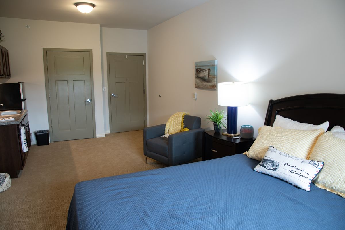 A cozy bedroom featuring a bed with blue bedding, an armchair, and a nightstand with a lamp.