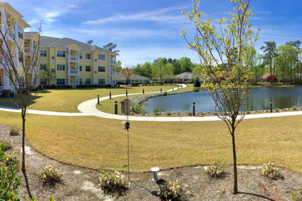 A serene view of an apartment complex with a pond and landscaped grounds under a clear blue sky.