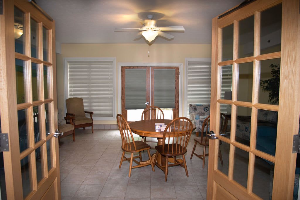A cozy dining room with a wooden table and chairs, featuring large windows and a ceiling fan.