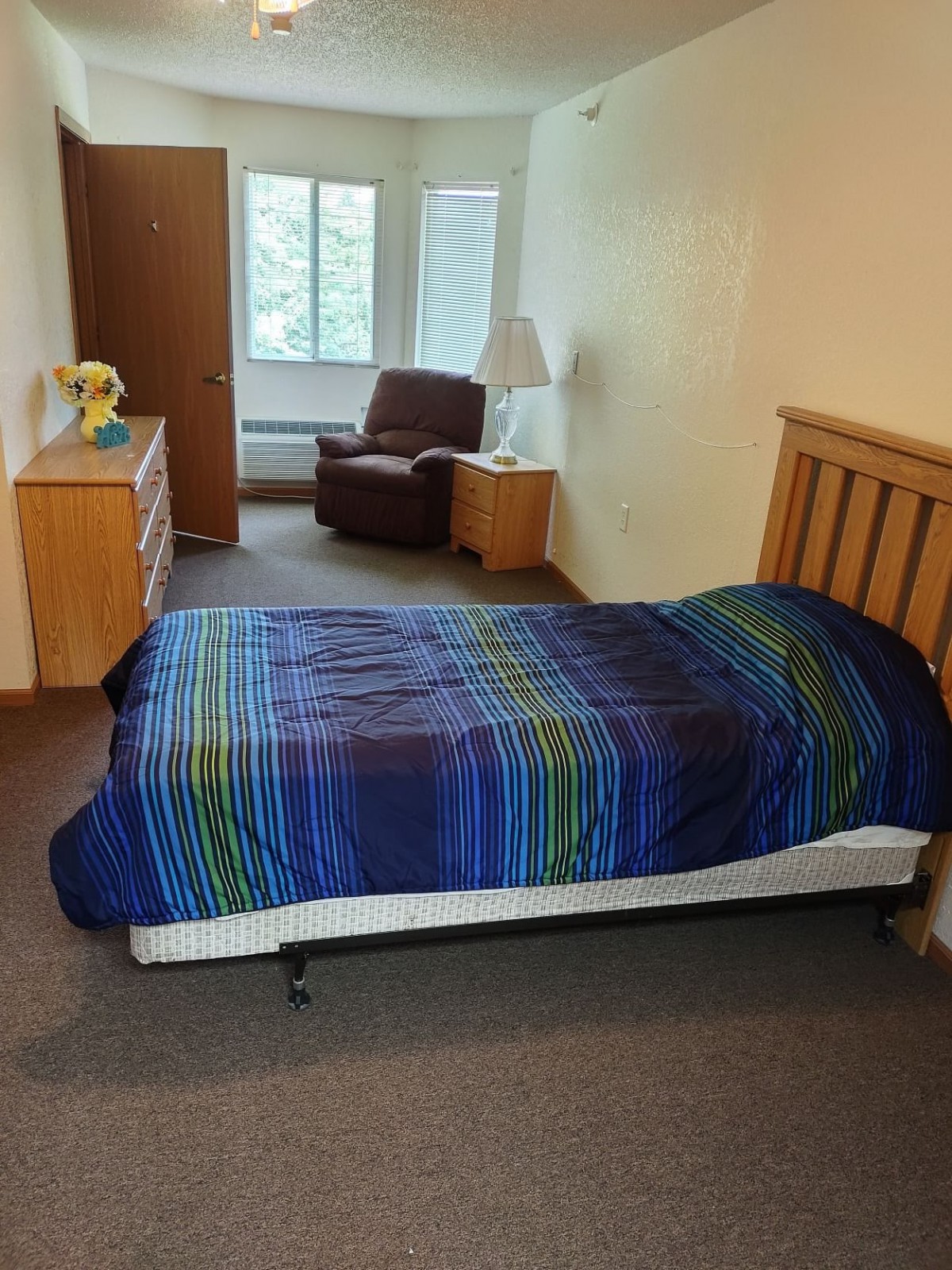 A cozy bedroom with a single bed, chair, and dresser, featuring a bright window.