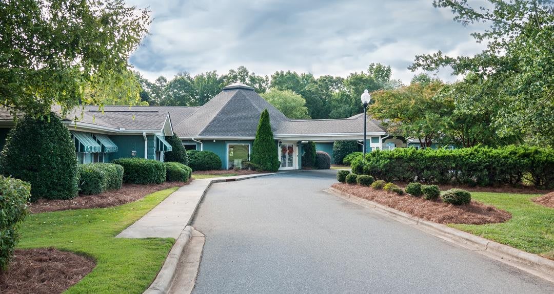 A view of a building with a circular driveway, surrounded by trees and landscaped gardens under a cloudy sky.