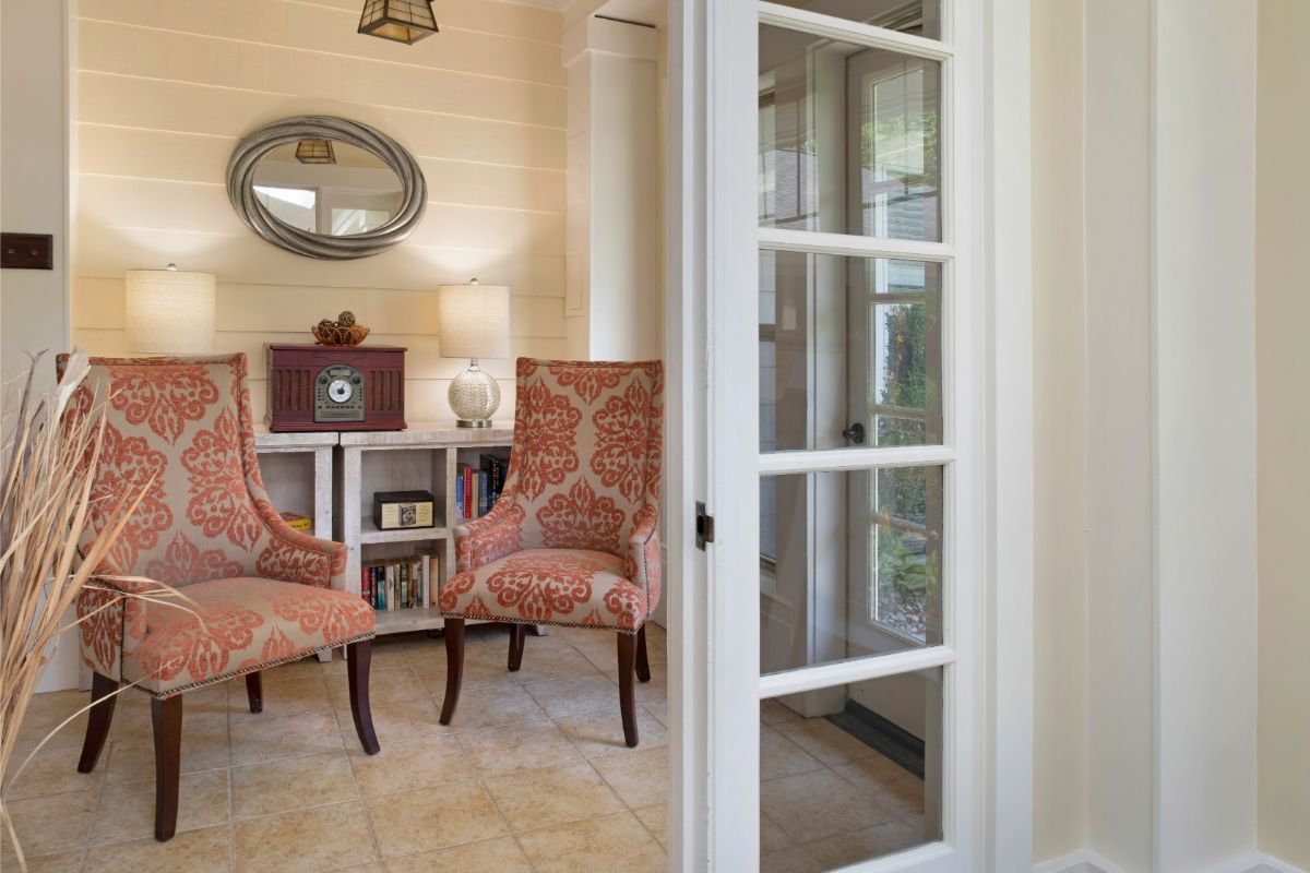 A cozy living room with two upholstered chairs and a round mirror, separated by a glass-paneled door.