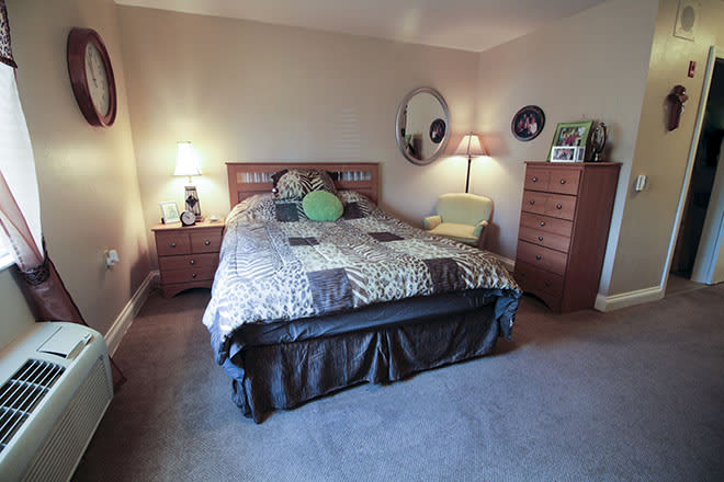 A cozy bedroom featuring a large bed with multiple pillows, two wooden dressers, lamps on either side of the bed, a round mirror on the wall, and carpeted flooring.