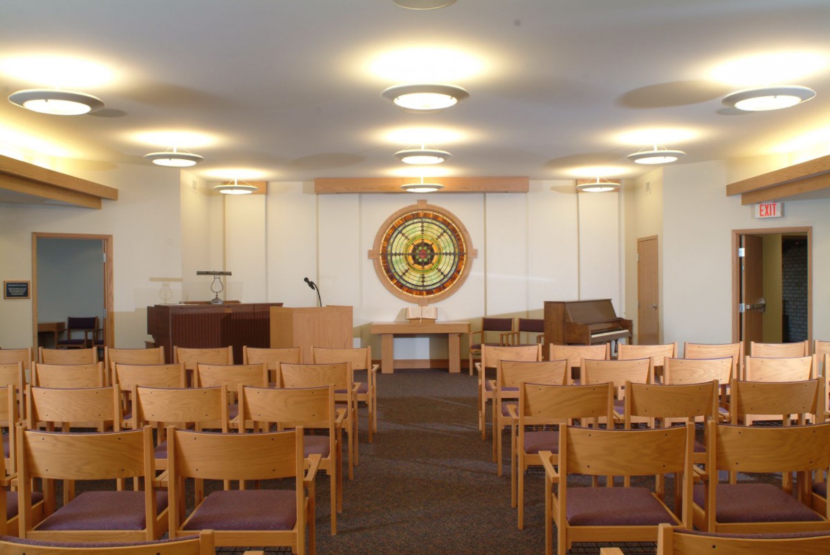 An interior view of a small chapel with wooden chairs and a stained glass window.