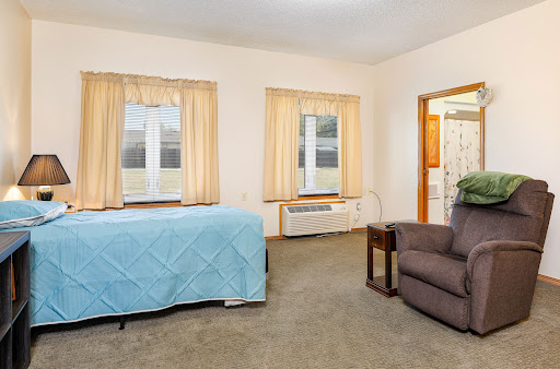 A cozy room featuring a bed with a blue coverlet, a comfortable chair, and large windows with light curtains.