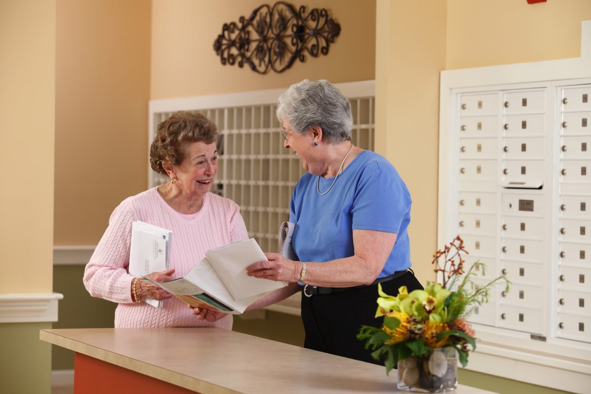 Two senior women engaged in a conversation at a reception desk, looking at paperwork together.